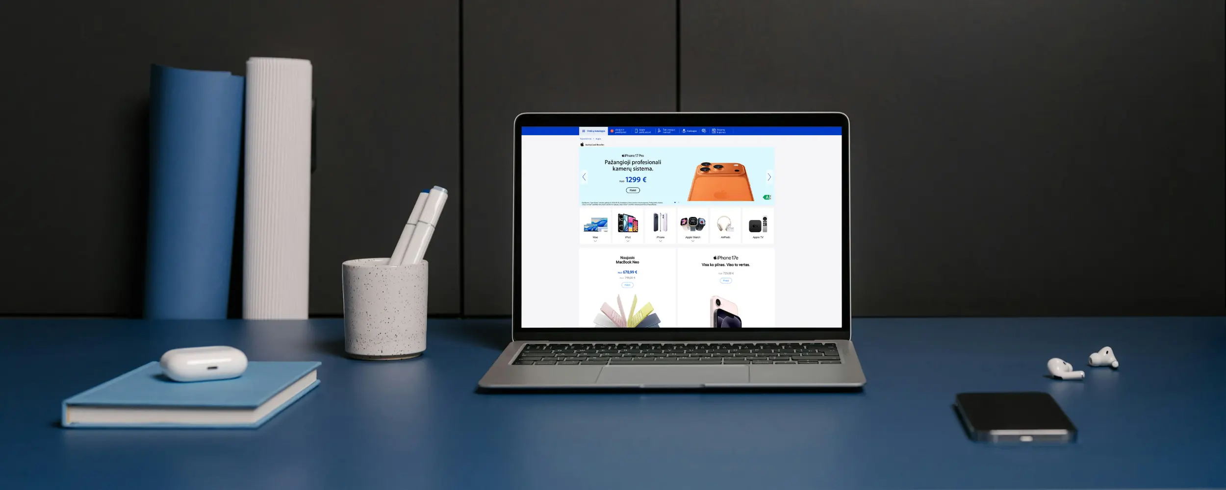 Laptop on a blue desk showing an online store page, with a notebook and wireless earbuds nearby in a tidy workspace.